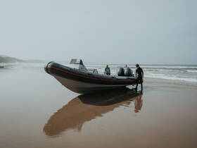 Two individuals prepare to embark on an adventure from a sleek boat on the beach, with the ocean waves gently lapping at the shore — a perfect setting for water sports or exploration.