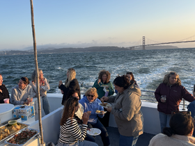 A lively group enjoys a sunset cruise, sharing food and drinks aboard a boat with the iconic Golden Gate Bridge in the background.