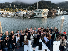 A vibrant group of women gathers on a boat, enjoying a festive celebration with drinks and smiles, set against a picturesque marina backdrop.