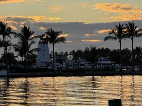 A picturesque sunset view featuring a lighthouse and marina, surrounded by palm trees, creating a serene atmosphere perfect for evening boat trips.