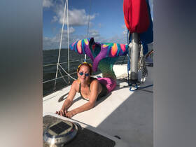 A woman dressed as a colorful mermaid poses on the deck of a sailboat, capturing the playful spirit of a fun day on the water.