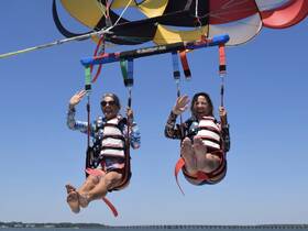 Two friends enjoy an exhilarating parasailing adventure, soaring high above the water with smiles and waves — a perfect way to experience the thrill of the ocean.