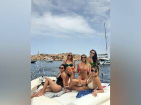 A group of friends enjoys cocktails on the deck of a yacht, celebrating summer in style against a scenic backdrop of rocky shores.