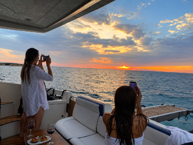 Two women capture the stunning sunset from the deck of a yacht, enjoying a picturesque moment with drinks and appetizers — perfect for memorable evening cruises.