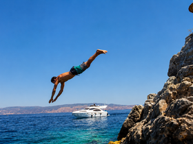 A man leaps off a rocky ledge into the clear blue waters, with a boat anchored nearby, capturing the essence of summer fun and adventure on the water.