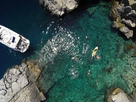 A vibrant scene of people swimming and kayaking in crystal-clear waters near a rocky coastline, with a boat anchored nearby — perfect for adventure seekers and water enthusiasts.