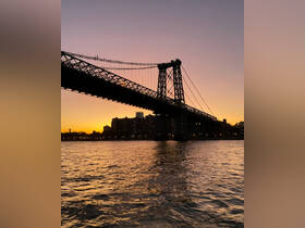 A stunning sunset view of the Williamsburg Bridge silhouetted against the city skyline, reflecting beautifully on the water — a perfect moment for evening boat tours.