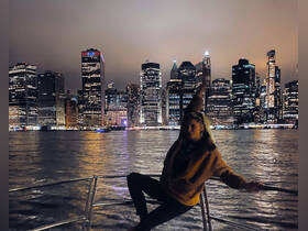 A woman poses on the bow of a boat against the stunning backdrop of a city skyline illuminated at night, capturing the essence of a vibrant evening cruise.
