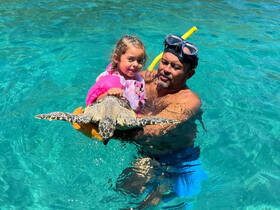 A child joyfully interacts with a sea turtle while being held by an adult in clear turquoise waters — a memorable wildlife experience perfect for family adventures.