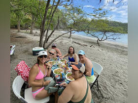 A lively group of friends enjoys a beachside meal, celebrating together with delicious food and drinks in a picturesque setting.