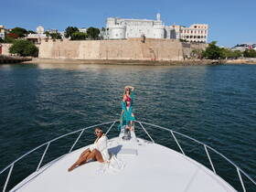 Two women pose stylishly on the bow of a yacht, with a historic fort and vibrant cityscape in the background — a perfect snapshot of a luxurious day on the water.