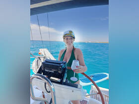 A smiling woman in a captain's hat enjoys a cocktail while steering a sailboat, capturing the essence of a fun sailing adventure on clear blue waters.