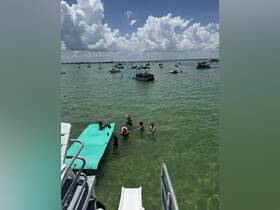 A lively group enjoys a sandbar stop, swimming and socializing in the clear waters, with multiple boats in the background — perfect for a fun day on the water.