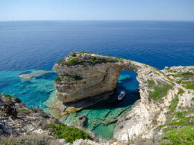 A stunning aerial view of a boat navigating through a natural rock arch along a picturesque coastline, showcasing the beauty of the surrounding turquoise waters.