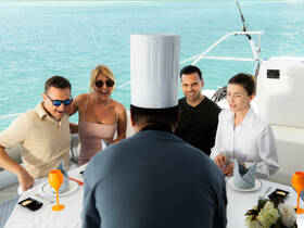 A lively group enjoys a gourmet dining experience aboard a yacht, with a chef presenting their meal against a stunning turquoise backdrop.