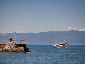 A couple enjoys the view from a pier as a boat sails by, showcasing the serene beauty of the waterfront and surrounding mountains.