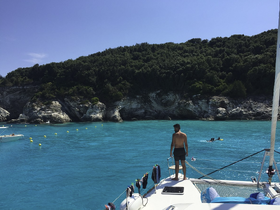 A person stands on the deck of a catamaran, overlooking a vibrant turquoise bay where others are enjoying swimming and snorkeling — a perfect day for water activities.