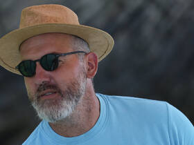 A man wearing sunglasses and a straw hat, captured in a candid moment. The background is not clearly defined, making the context ambiguous.