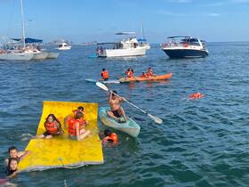 A lively group enjoys a fun day on the water, featuring kayaking, swimming, and relaxing on a floating mat, all while wearing life jackets for safety.