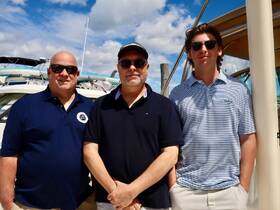 Three men pose together at a marina, showcasing a sunny day and a backdrop of boats — perfect for capturing memories before a day on the water.