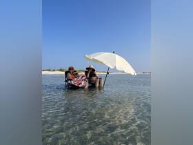 Two friends relax in shallow water under a large umbrella, enjoying a sunny beach day — a perfect setting for unwinding and soaking up the sun.
