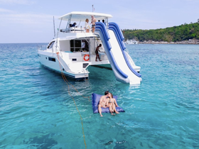 A couple enjoys a romantic moment on a floating mat in crystal-clear waters, with a fun boat slide in the background — perfect for adventurous getaways.