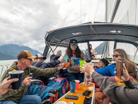 A lively group aboard a sailboat raises their drinks in celebration, enjoying a memorable sailing experience surrounded by stunning mountain views.