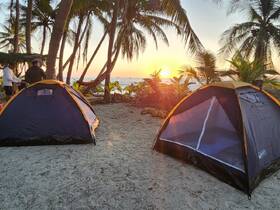 A serene beach scene at sunset featuring tents and palm trees, with individuals preparing for a camping experience by the water.