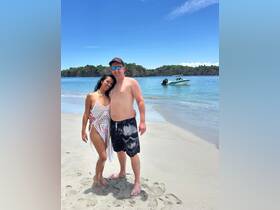 A couple poses together on the beach with a boat in the background, capturing a perfect moment of their sunny vacation by the water.