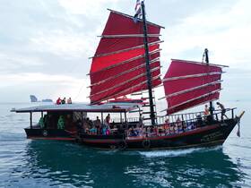 A vibrant group enjoys a sailing experience aboard a traditional boat with striking red sails, set against a scenic backdrop of calm waters and distant islands.