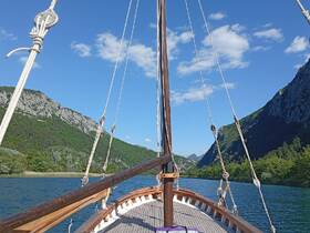 A scenic view from the front of a sailboat navigating through a tranquil waterway, surrounded by lush green mountains and a clear blue sky.