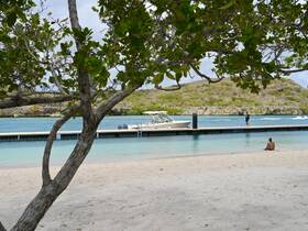 A serene beach scene featuring a boat docked at a pier, with a person relaxing on the sand and another enjoying the view — perfect for a day of leisure by the water.