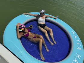 Two friends relax on a floating mat in the water, enjoying drinks and sunshine — a perfect way to unwind during a summer day on the water.