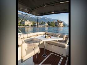 A beautifully arranged interior of a boat featuring a dining area with wine and snacks, set against a stunning lakeside backdrop of mountains and a castle.