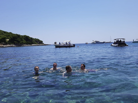 A lively group enjoys a refreshing swim in crystal-clear waters, with boats anchored nearby, perfect for a fun day at the beach.