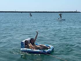 Two friends enjoy a sunny day floating on a mat while paddleboarders glide by in the background — a perfect scene for summer fun on the water.