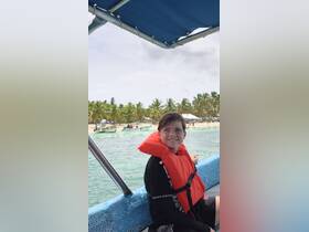A young boy wearing a life jacket smiles while aboard a boat, with a beautiful beach and palm trees in the background — perfect for family adventures on the water.