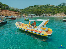 A vibrant boat named Vagabundo navigates through crystal-clear waters near a bustling beach, perfect for sea safari adventures and summer fun.