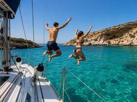 A couple joyfully jumps off the side of a sailboat into the crystal-clear turquoise waters, capturing the essence of summer fun and adventure on the sea.