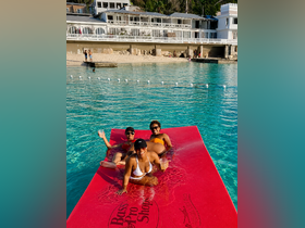 A fun group enjoys drinks on a floating mat in crystal-clear waters, perfect for a relaxing day at the beach or poolside party.