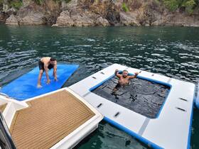 Two friends enjoy a fun day on the water, one balancing on a floating mat while the other relaxes in a hot tub — a perfect way to unwind and enjoy the sun.