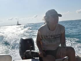 A woman enjoys a sunny day on the water, steering a boat with a sailboat visible in the background — perfect for adventure seekers and sailing enthusiasts.