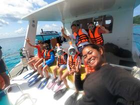 A joyful group of snorkelers, including children, poses for a selfie on a boat, ready for an exciting underwater adventure in crystal-clear waters.