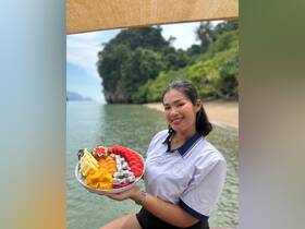 A smiling staff member presents a vibrant fruit platter on a serene beach, perfect for catering on boat trips or beach outings.