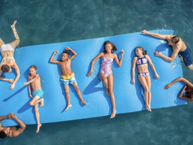 A lively group of children and adults enjoy a sunny day on a floating mat in clear waters, showcasing the perfect summer fun experience.