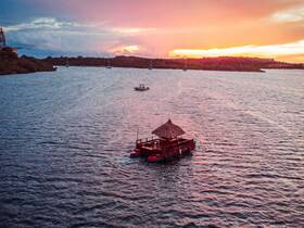 A picturesque aerial view of a tiki boat anchored in calm waters during sunset, surrounded by lush greenery and distant sailboats, perfect for a serene getaway.
