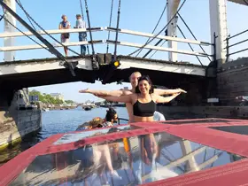 A joyful couple poses with arms outstretched on a boat as they pass under a bridge, capturing the essence of a fun day on the water.