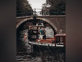 A couple poses on a charming boat in Amsterdam's canals, with a picturesque bridge in the background — a perfect moment capturing the romance of city exploration.