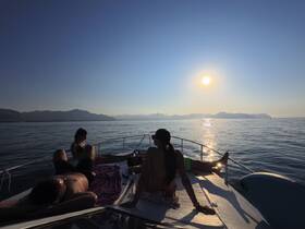 A group of friends relaxes on the deck of a boat during a stunning sunset, enjoying the serene waters and beautiful mountain backdrop — perfect for a memorable evening on the water.