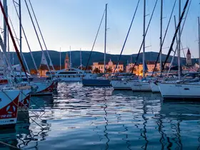 A serene evening view of various yachts and sailboats docked in a picturesque harbor, reflecting the warm lights of the waterfront buildings.
